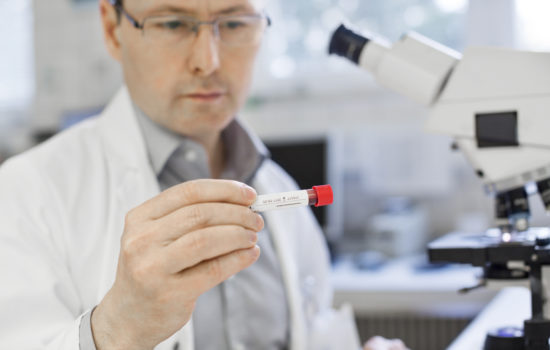 Male chemist analyzing at sample in test tube at laboratory. Scientist is sitting at table in hospital. He is in lab coat.