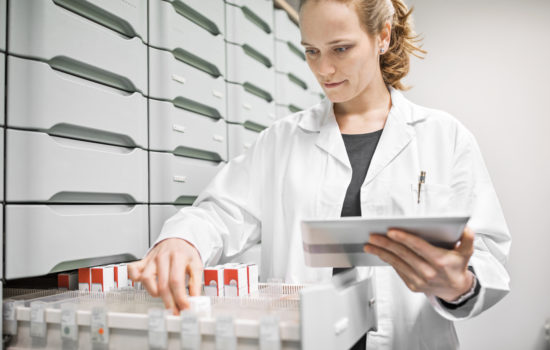 Female chemist searching for medicines in drawer. Pharmacist is holding digital tablet while working in storage room. She is wearing lab coat at pharmacy.